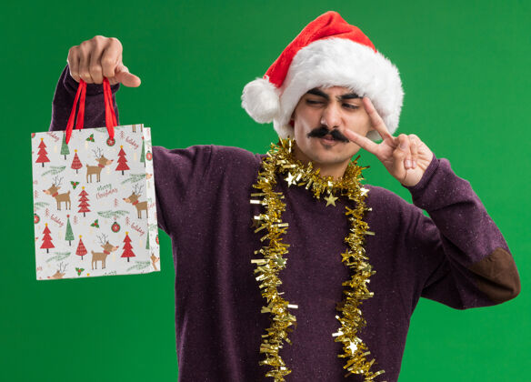 圣诞老人年轻的胡子男人戴着圣诞老人帽子 脖子上挂着金属丝 手里拿着圣诞礼物的纸袋 满脸喜悦地看着它 绿色的背景上立着v字标志年轻圣诞快乐快乐