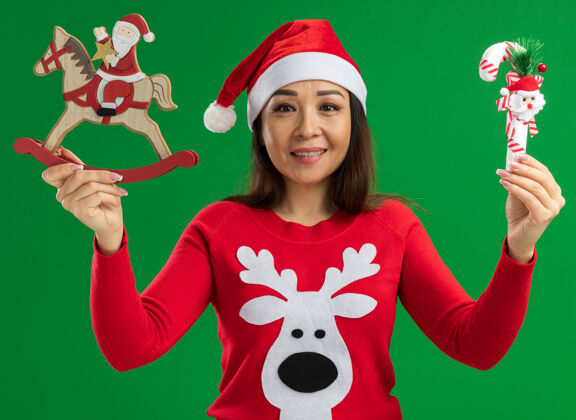 拿着快乐的年轻女子戴着圣诞老人帽 穿着红色毛衣 拿着圣诞玩具 面带微笑地看着相机 站在绿色的背景下脸微笑圣诞快乐
