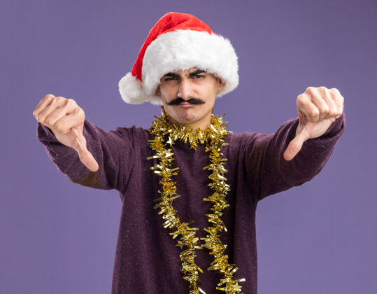 金属片年轻的胡子男人戴着圣诞老人帽子 脖子上戴着金属丝 看着摄像机不高兴地竖起大拇指站在紫色的背景上周围圣诞老人圣诞
