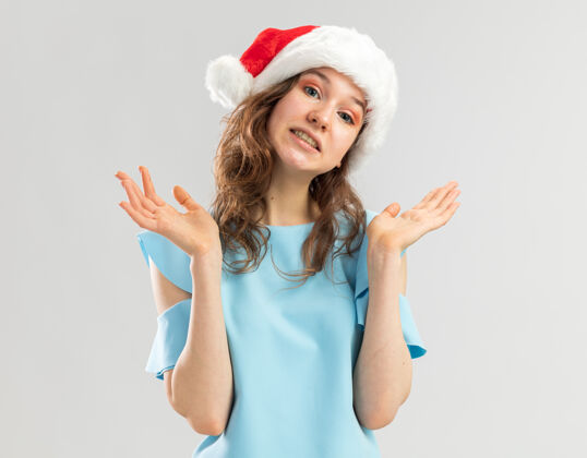 圣诞快乐戴着蓝色上衣和圣诞帽的年轻女子看起来很困惑 举起双臂圣诞老人困惑举起