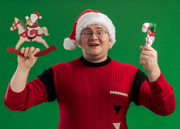 圣诞老人快乐的成年男子戴着眼镜和圣诞帽 拿着摇摆木马上的圣诞老人雕像和糖果手杖饰品 看着隔离在绿色背景上的相机举行马雕像