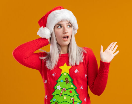 金发穿着圣诞毛衣 戴圣诞帽的金发女郎站在橙色的背景上 抬起手臂 看起来很惊讶橙色圣诞老人抬高