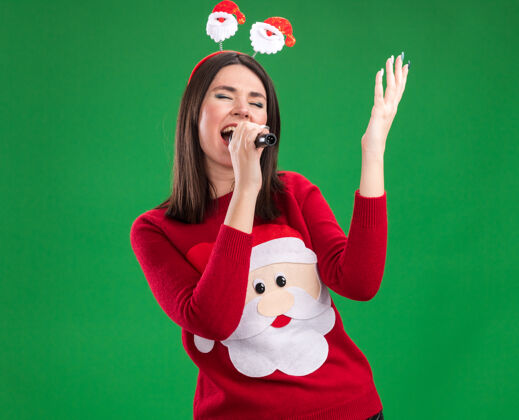 年轻年轻漂亮的白人女孩穿着圣诞老人的毛衣和头带拿着麦克风举起手唱歌闭着眼睛隔离在绿色的墙壁与复制空间圣诞老人穿头带