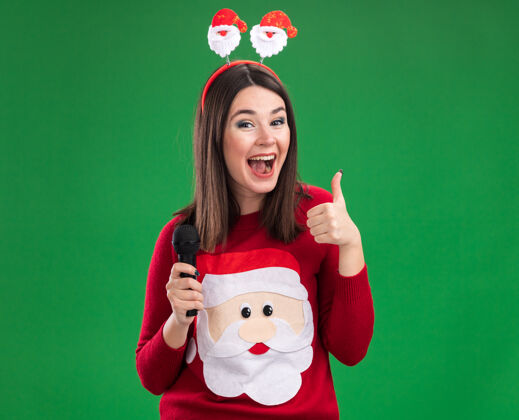 展示令人印象深刻的年轻漂亮的白人女孩穿着圣诞老人毛衣和头带拿着麦克风显示拇指向上隔离在绿色墙上的复制空间麦克风毛衣圣诞老人