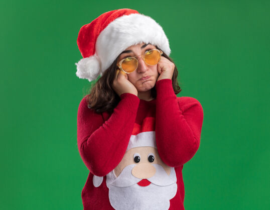 圣诞老人穿着圣诞毛衣 戴圣诞帽 戴圣诞眼镜的年轻女孩站在绿色的墙上 神情悲伤地看着一边悲伤年轻站在一边