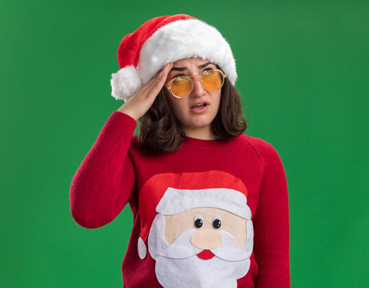 年轻穿着圣诞毛衣 戴着圣诞帽和眼镜的年轻女孩站在绿色的墙上 眼睛直打转 又累又无聊圣诞节累了无聊