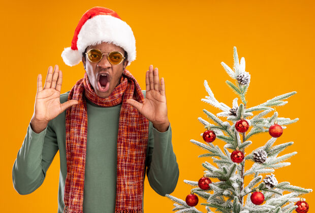圣诞老人戴着圣诞帽 脖子上围着围巾的非裔美国人站在橙色墙上的圣诞树旁 带着咄咄逼人的表情大喊大叫喊叫男人围巾