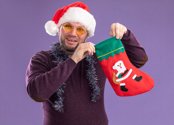 长袜快乐的中年男子戴着圣诞帽 脖子上戴着金箔花环 戴着眼镜 手里拿着圣诞长袜 孤零零地站在紫色的墙上圣诞老人周围男人