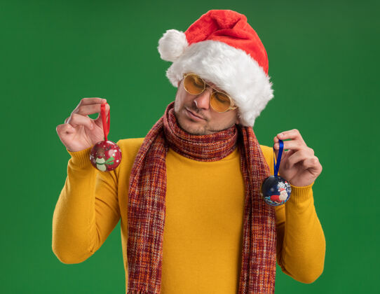 高领毛衣一个穿着黄色高领毛衣 戴着圣诞帽 戴着眼镜 拿着圣诞树玩具的年轻人站在绿色的墙上严肃地看着他们圣诞节年轻男人