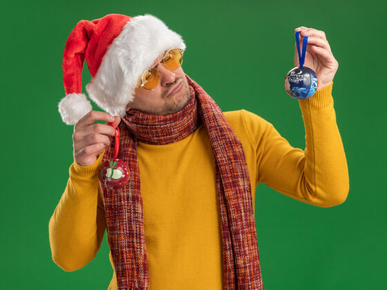 站立一个穿着黄色高领毛衣 戴着圣诞帽 戴着眼镜 拿着圣诞树玩具的年轻人站在绿色的墙上 困惑地看着玩具 满脸疑惑疑惑玩具年轻人