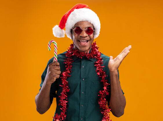 高兴快乐的非洲裔美国人戴着圣诞帽 戴着花环 戴着眼镜 拿着糖果手杖 高高兴兴地微笑着 手臂举过橙色的墙花环站着帽子