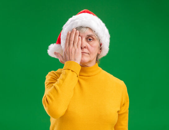 手自信的老妇人戴着圣诞老人的帽子 用手遮住眼睛 在紫色的背景上与复制空间隔离帽子眼睛自信