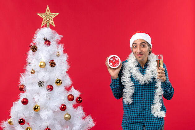 帽子带着圣诞老人帽子 举着一杯葡萄酒 拿着一个站在圣诞树旁的闹钟 满脸笑容 情绪激动的年轻人站着葡萄酒玻璃