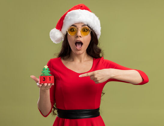 圣诞树年轻漂亮的女孩戴着圣诞帽 戴着眼镜 拿着圣诞树玩具 看着约会年轻女孩日期