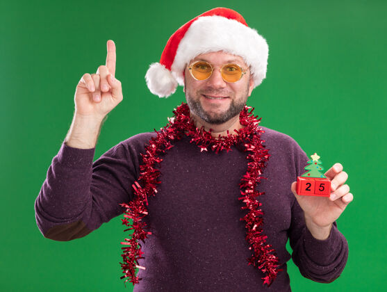 脖子高兴的中年男子戴着圣诞帽 脖子上戴着金箔花环 戴着眼镜 手里拿着圣诞树玩具和日期相机举行看