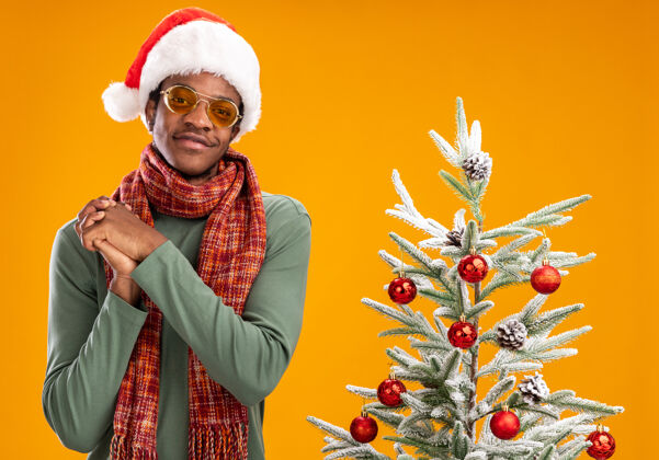 微笑戴着圣诞帽 脖子上围着围巾的非洲裔美国人看着镜头 幸福而积极地微笑着 手牵着手站在橙色背景下的圣诞树旁相机手男人