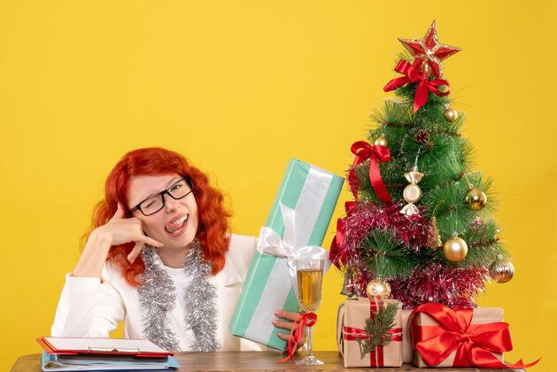 坐着正面图：女医生坐在黄色的桌子上 手里拿着圣诞礼物和圣诞树情感安排树