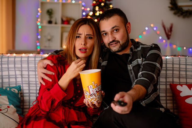 装饰一对年轻漂亮的夫妇惊讶地坐在沙发上拿着一桶爆米花的女人和男人在装饰好的房间里一起看电视 背景是圣诞树圣诞年轻沙发