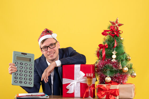 计算器商务人士手持计算器坐在圣诞树旁的桌子前 呈现黄色男桌子圣诞节