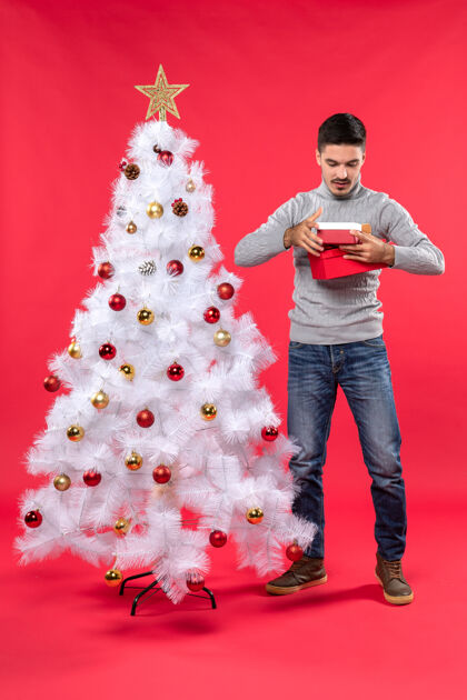 雪人一个穿着灰色上衣的浪漫成年人站在装饰过的白色圣诞树旁 手里拿着红色的礼物礼物衬衫欢快