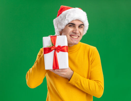 站着穿着黄色高领毛衣 戴着圣诞老人帽的快乐的年轻人拿着礼物 站在绿色的背景下 兴高采烈地微笑着看着相机帽子微笑年轻人