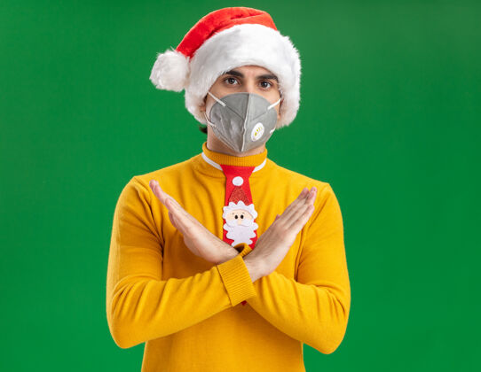 手穿着黄色高领毛衣 戴着圣诞老人帽 戴着滑稽的领带 戴着面罩 做着停下来的手势 双手交叉看着相机 严肃的脸站在绿色的背景上交叉高领毛衣相机