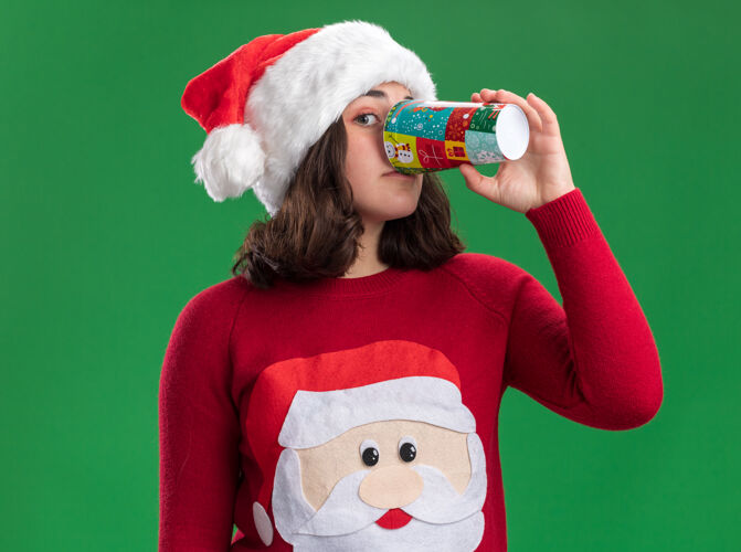 圣诞老人穿着圣诞毛衣 戴着圣诞帽的小女孩站在绿色的墙上 从五颜六色的纸杯里喝水站着喝酒年轻人