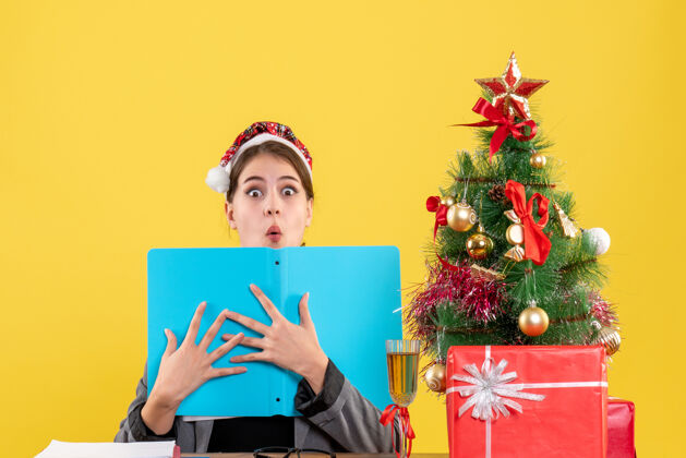 手前视图震惊的女孩戴着圣诞帽坐在桌旁 双手拿着文件夹圣诞树和礼物鸡尾酒桌子文件夹文件