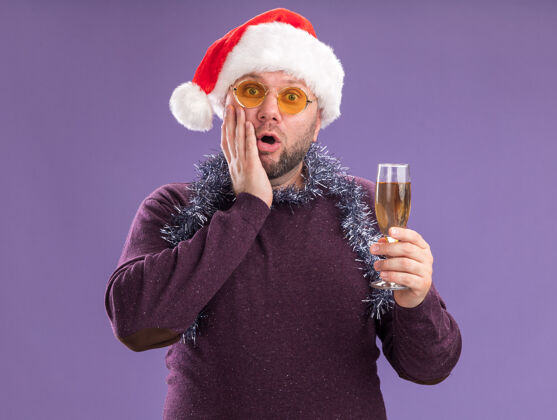 圣诞老人关心的中年男子戴着圣诞帽 脖子上戴着金箔花环 戴着眼镜 手里拿着香槟酒杯 手放在脸上 隔离在紫色的墙上花环帽子保管
