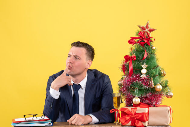 圣诞西装革履的男人把手指放在嘴边坐在圣诞树旁的桌子上 黄色的礼物手指穿西装的男人西装
