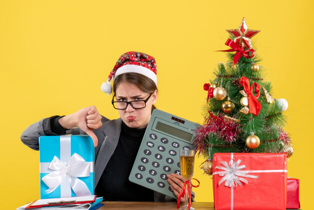 标志前视图悲伤的女孩戴着圣诞帽坐在桌子旁做拇指向下的标志圣诞树和礼物鸡尾酒树圣诞节人