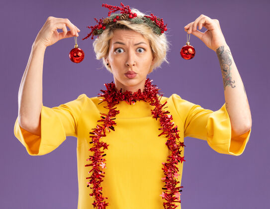圣诞饰品笨拙的金发女郎头戴圣诞花环 脖子上戴着金箔花环 手拿圣诞饰品 头靠紫色墙上的隔离墙圣诞节头附近