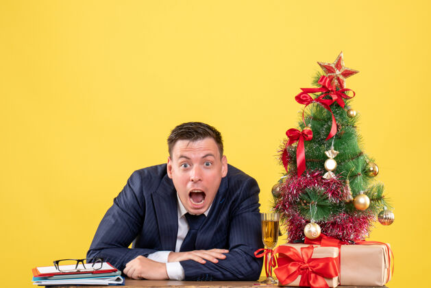 商人正面图惊奇的人张开嘴坐在圣诞树旁的桌子旁 黄色的墙上摆着礼物树嘴西装