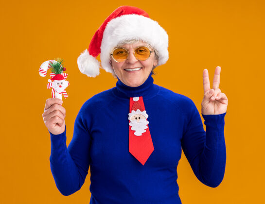 胜利戴着太阳眼镜 戴着圣诞帽 系着圣诞领带 面带微笑的老妇人手持糖果手杖 在橙色背景上用复制空间孤立地示意胜利标志举行糖果眼镜
