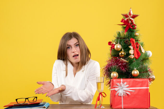 快乐正面图：女医生坐在黄色背景的桌子前 放着圣诞树和礼品盒健康圣诞节女性