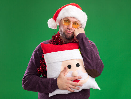 枕头无聊的中年男子戴着圣诞帽 脖子上戴着金丝花环 戴着眼镜 抱着圣诞老人的枕头 手放在脸上 隔离在绿色的墙上圣诞老人保管金属丝