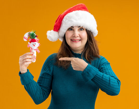 持有令人印象深刻的成年白人妇女与圣诞老人帽子举行和点糖果甘蔗孤立的橙色背景与复制空间帽子印象深刻手杖