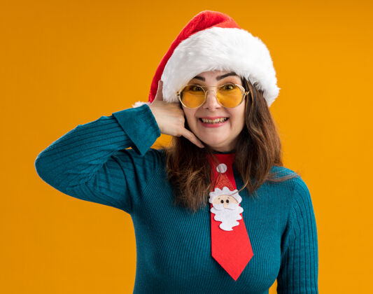 微笑戴着太阳眼镜 戴着圣诞帽 打着圣诞领带 面带微笑的成年白种女人在橙色背景上用复制空间孤立地打着“呼叫我”的手势电话手势眼镜