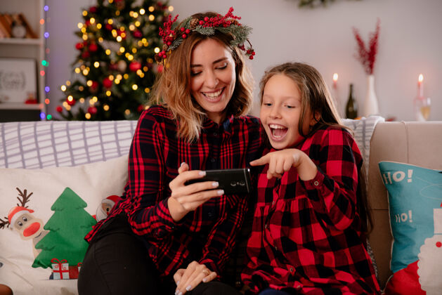 电话笑眯眯的母女俩坐在沙发上看电话 在家里享受圣诞节时光享受笑圣诞节