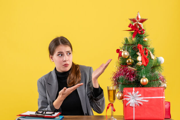 展示前视图惊讶的女孩坐在桌子旁展示圣诞树和鸡尾酒礼物成人坐着专业