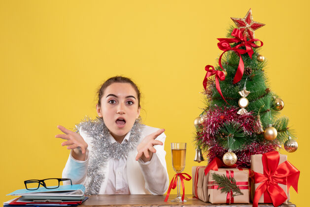 背景正面图：女医生坐在黄色背景的桌子后面 还有圣诞树和礼品盒圣诞节健康花