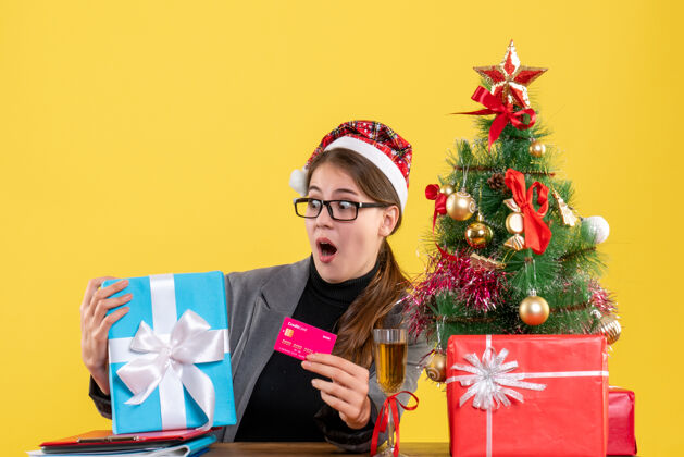 圣诞节正面图戴着圣诞帽的大眼睛女孩坐在桌子旁 手里拿着卡片圣诞树和鸡尾酒礼物礼物桌子幸福