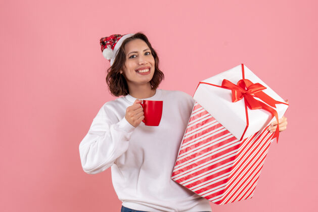 前面年轻女子手持圣诞礼物和粉红色墙上的一杯茶的前视图杯子礼物年轻