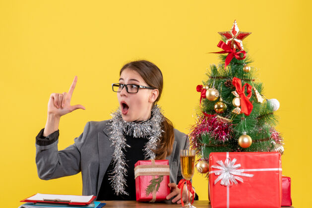 肖像前视图困惑的女孩戴着眼镜坐在桌边指着圣诞树和礼物鸡尾酒圣诞节桌子礼物