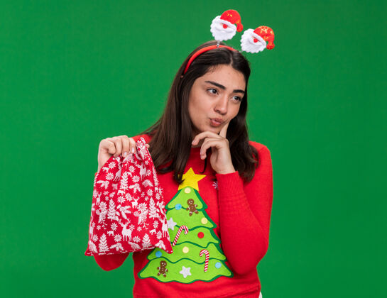 抱体贴的年轻白种女孩戴着圣诞老人的头带 手指放在下巴上 拿着圣诞礼品袋 看着绿色背景上孤立的一面 还有复制空间头带年轻放
