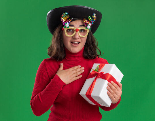 惊喜穿着红毛衣 戴着滑稽眼镜 戴着黑帽子 拿着礼物的年轻女孩站在绿色的墙上 惊喜而快乐圣诞节毛衣穿着