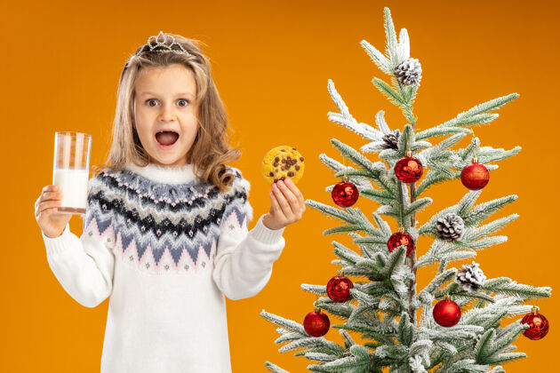 就在附近快乐的小女孩站在圣诞树旁 戴着头饰 脖子上戴着花环 手里拿着一杯牛奶 橘黄色的墙上隔着饼干快乐小戴着