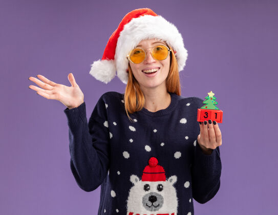 女孩微笑的年轻漂亮女孩穿着圣诞毛衣戴着圣诞帽戴着眼镜拿着圣诞玩具摊开双手孤立在紫色背景上毛衣手年轻
