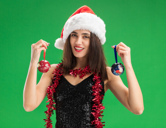 帽子微笑着的年轻漂亮女孩戴着圣诞帽 脖子上戴着花环 手里拿着圣诞树上的球 隔离在绿色的墙上女人脖子球