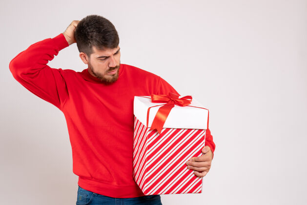 肖像身穿红衫的年轻人在白色墙上的盒子里拿着圣诞礼物前面微笑礼物